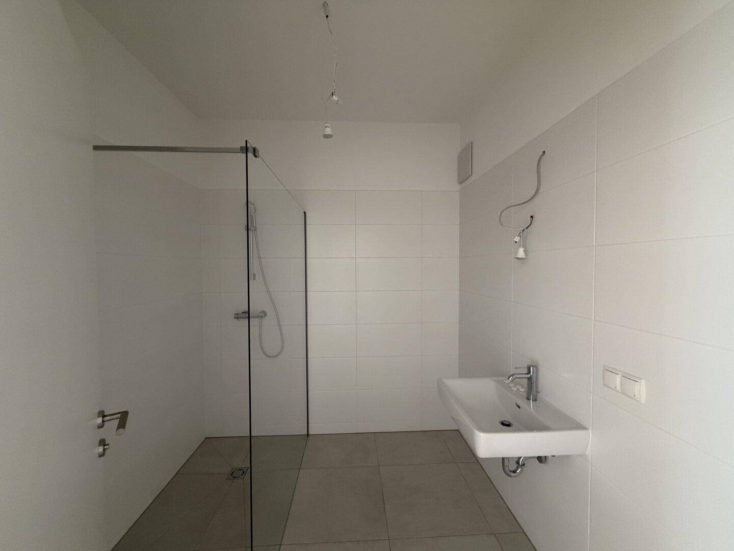 Modernes Badezimmer mit weißen Fliesen, begehbarer Dusche und wandmontiertem Waschbecken.