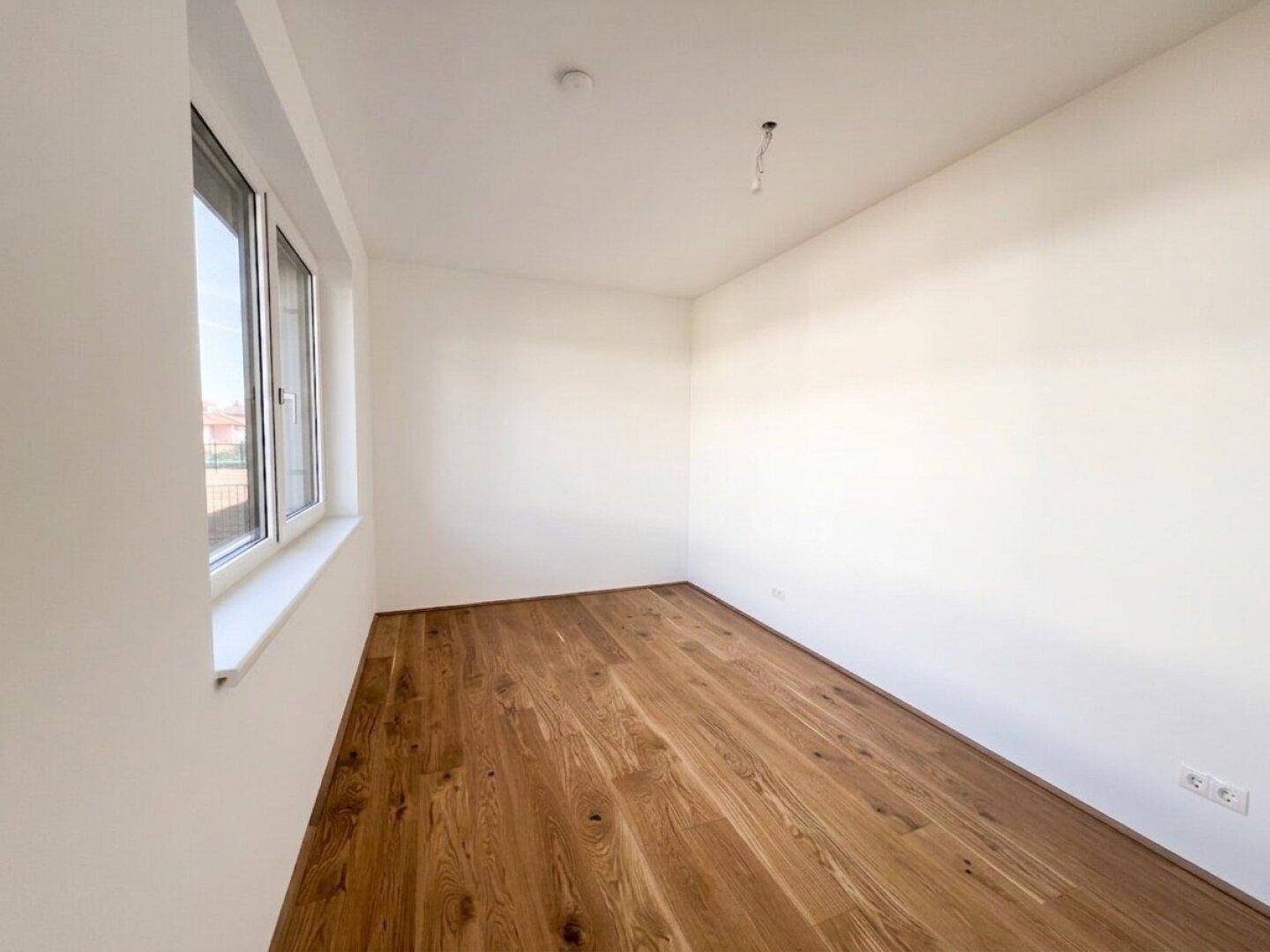 Leeres Schlafzimmer mit Holzboden und einem großen Fenster, das viel Tageslicht hereinlässt.
