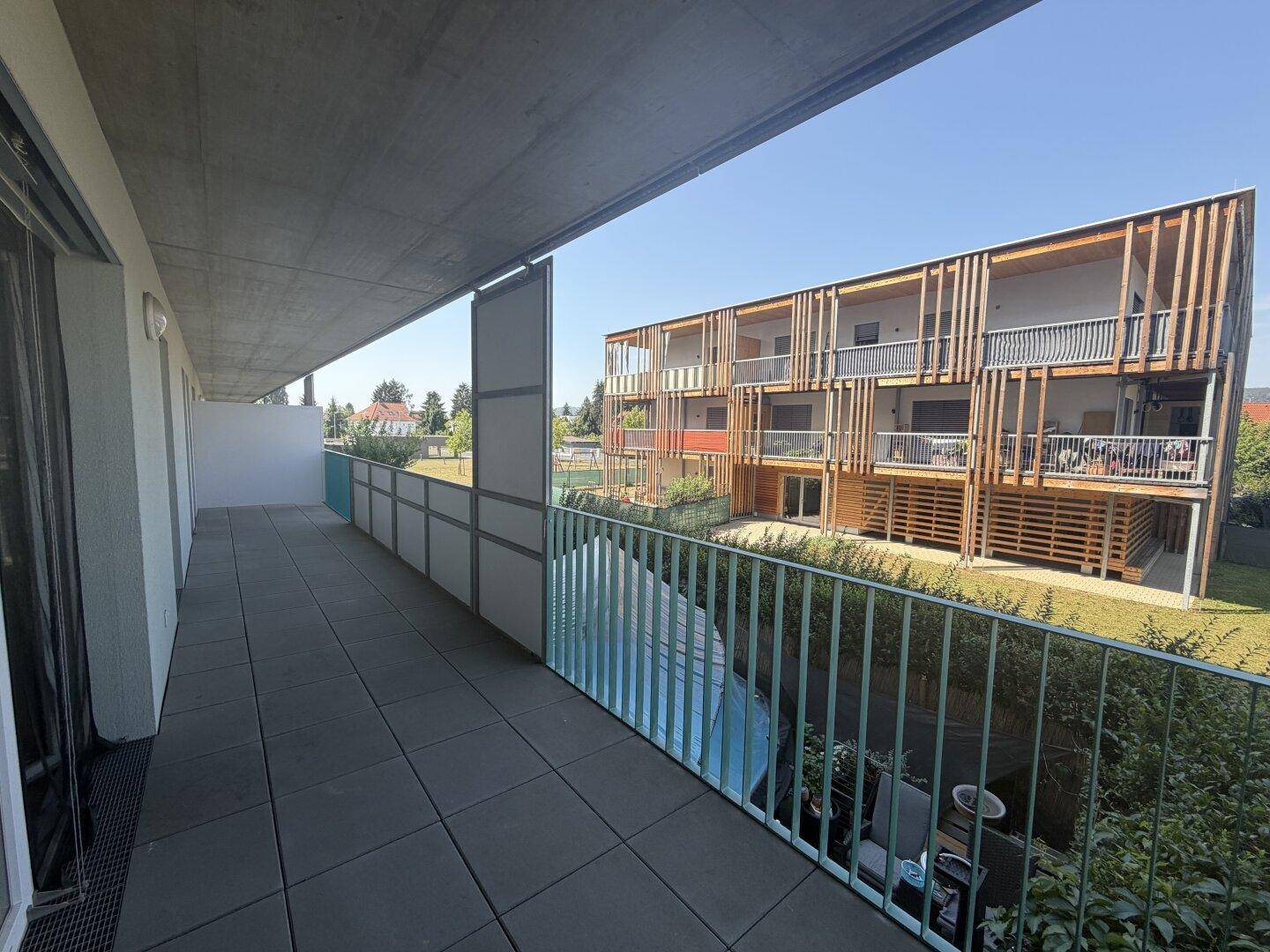 Langer Balkon mit Fliesenboden und Blick auf die moderne Architektur des Nachbargebäudes und Grünflächen.