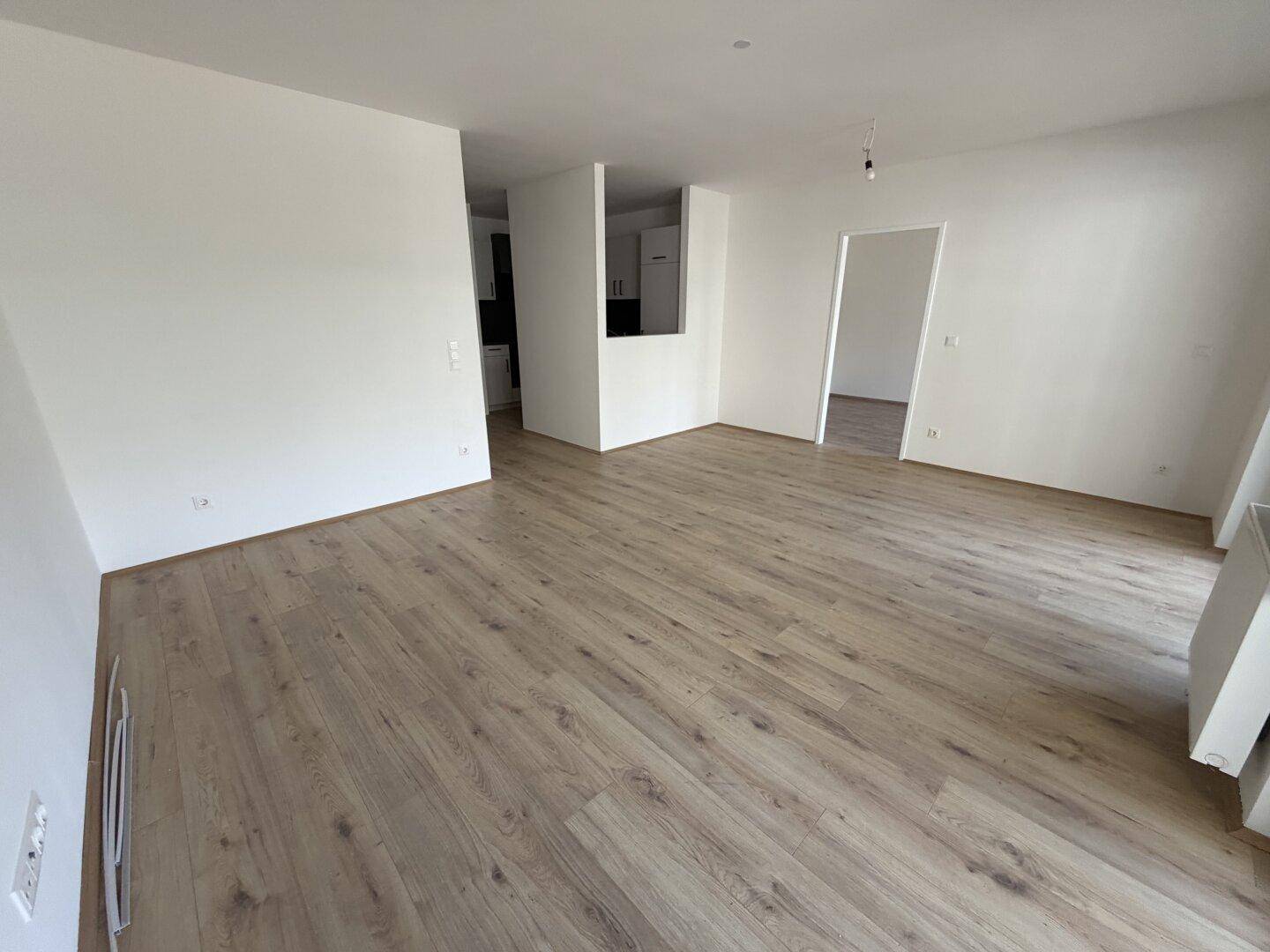 Geräumiger Wohnbereich mit hellem Holzboden und teilweisem Blick auf die moderne Küche.