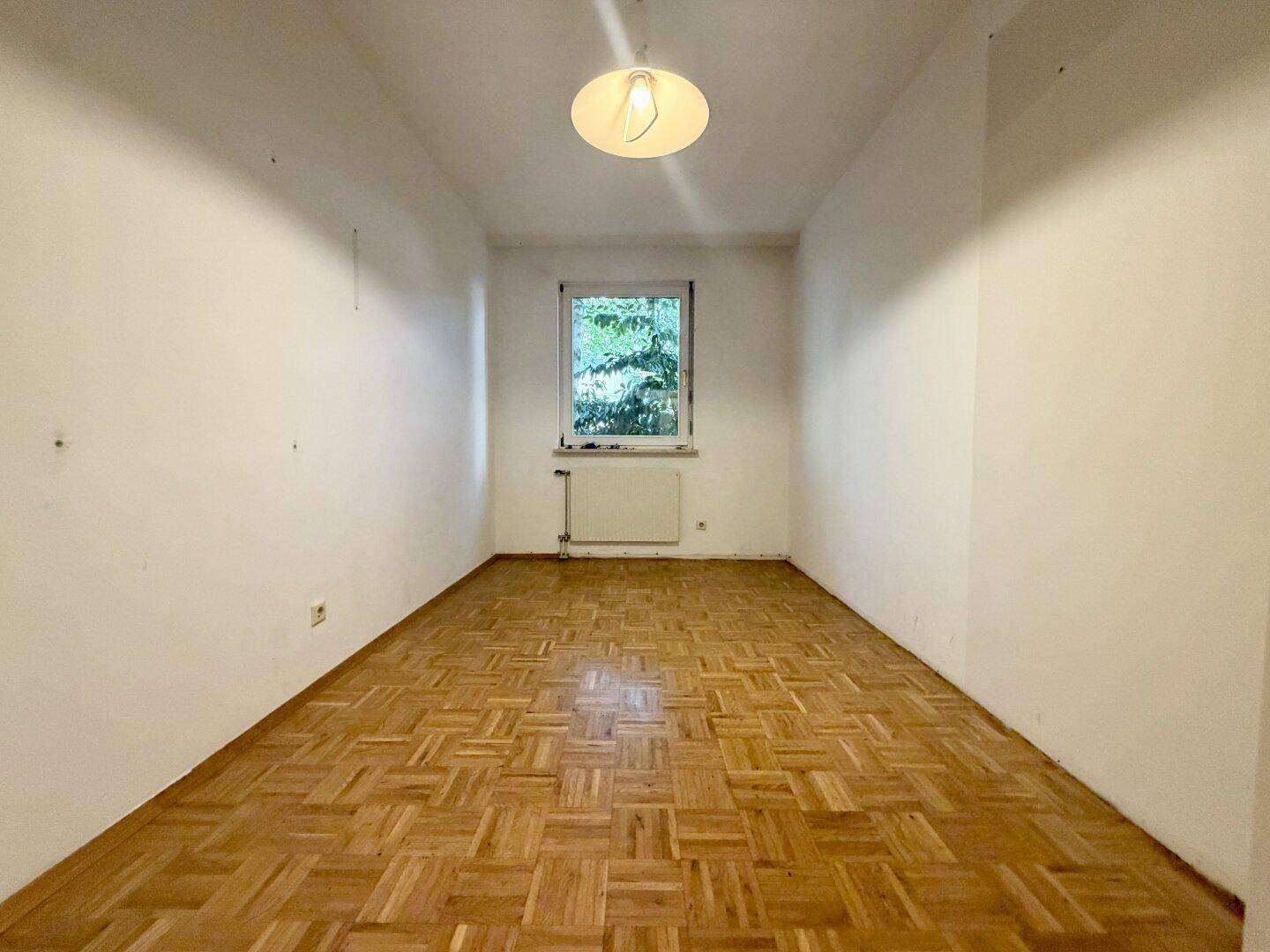 Leeres Zimmer mit Parkettboden und einem Fenster mit Blick ins Grüne.