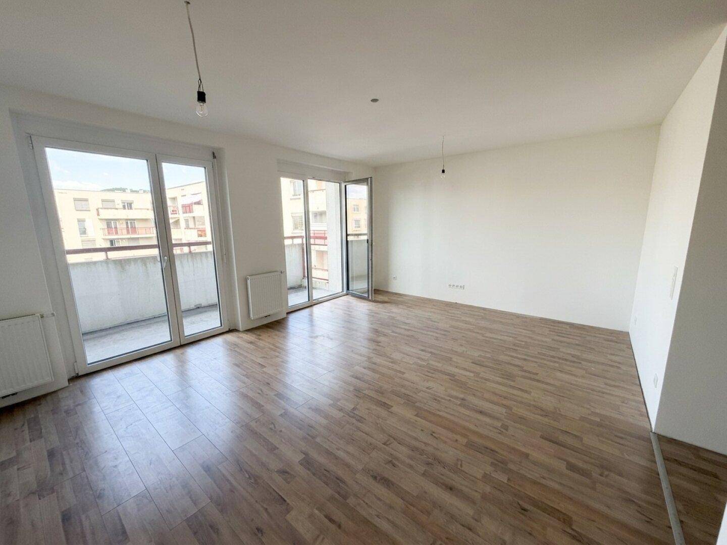 Geräumiges Wohnzimmer mit Laminatboden und großen Fenstern, die viel Tageslicht hereinlassen.