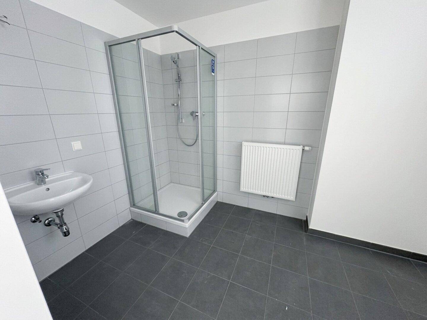 Helles Badezimmer mit grauen Fliesen, einer Duschkabine und einem Waschbecken.