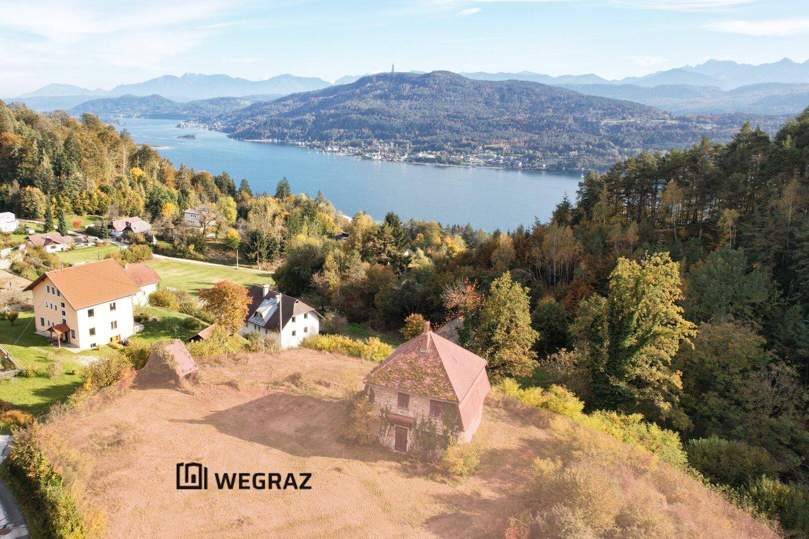 Luftaufnahme eines Grundstücks mit einem alten Haus, umgeben von Bäumen, mit einem See und Bergen im Hintergrund.