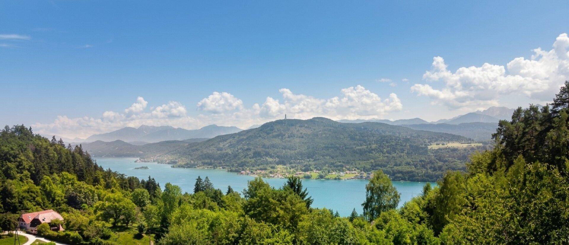 Panoramablick auf einen See umgeben von bewaldeten Hügeln und fernen Bergen unter blauem Himmel.