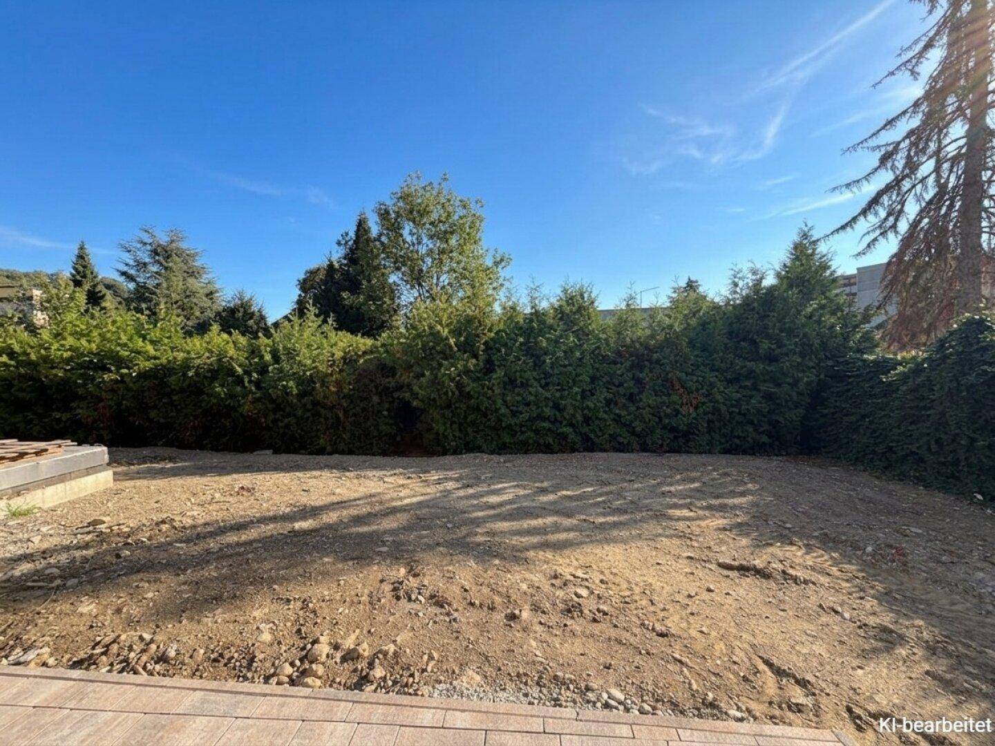 Unfertige Gartenfläche mit bloßer Erde und Hecken im Hintergrund unter einem klaren blauen Himmel.