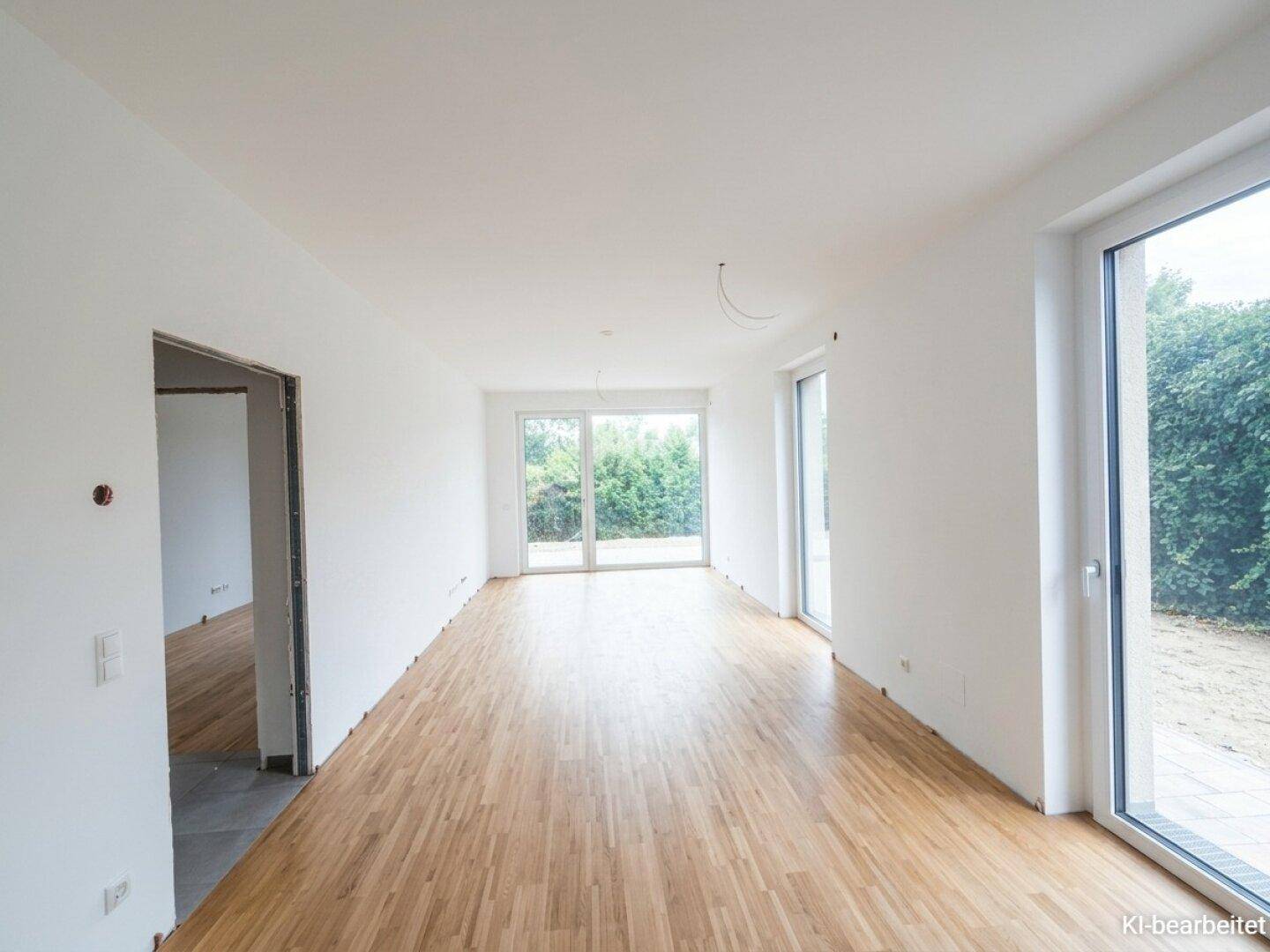 Geräumiger, unmöblierter Wohnbereich mit hellem Holzboden und Zugang zum Außenbereich durch große Fenster.