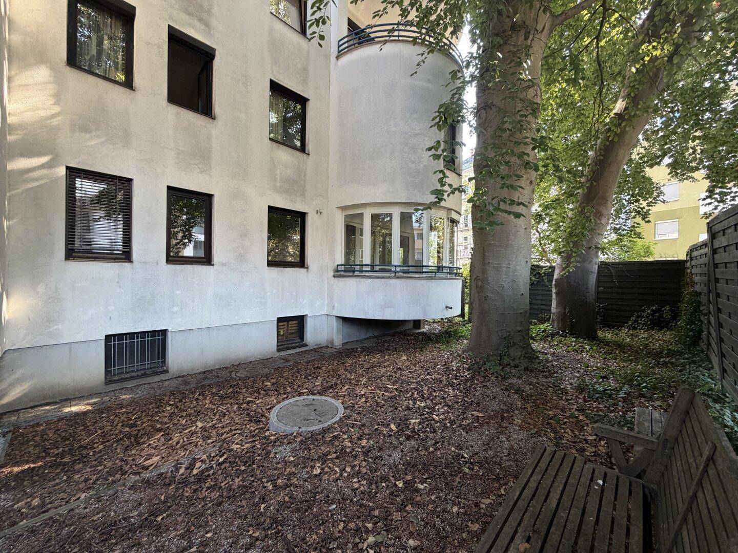 Seitenansicht des Gebäudes mit Gartenbereich, altem Baum und einer Holzbank im Vordergrund.