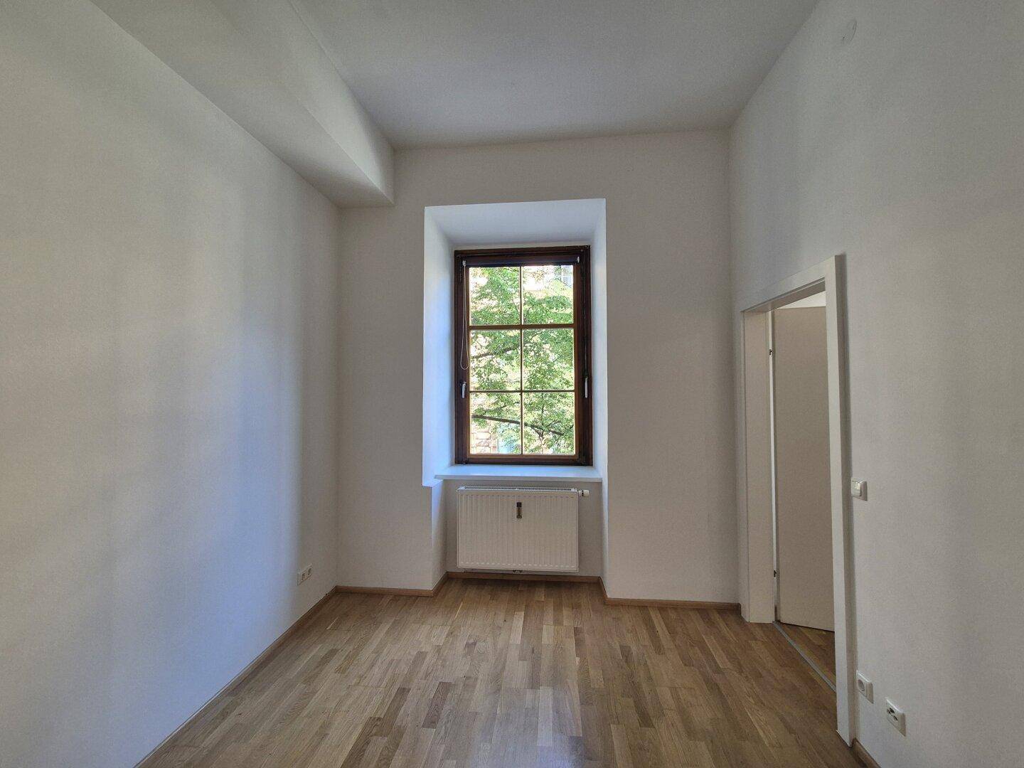 Helles Zimmer mit Parkettboden und einem großen Fenster, das viel Tageslicht hereinlässt.