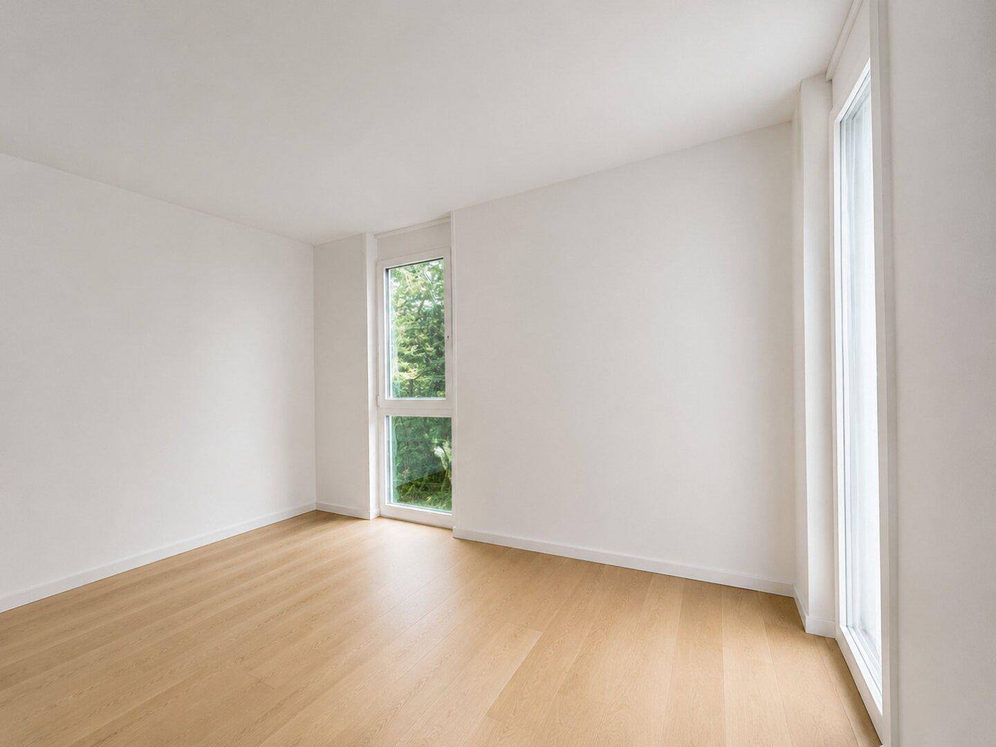 Leeres Schlafzimmer mit Holzboden und hohen Fenstern, die viel Tageslicht hereinlassen.