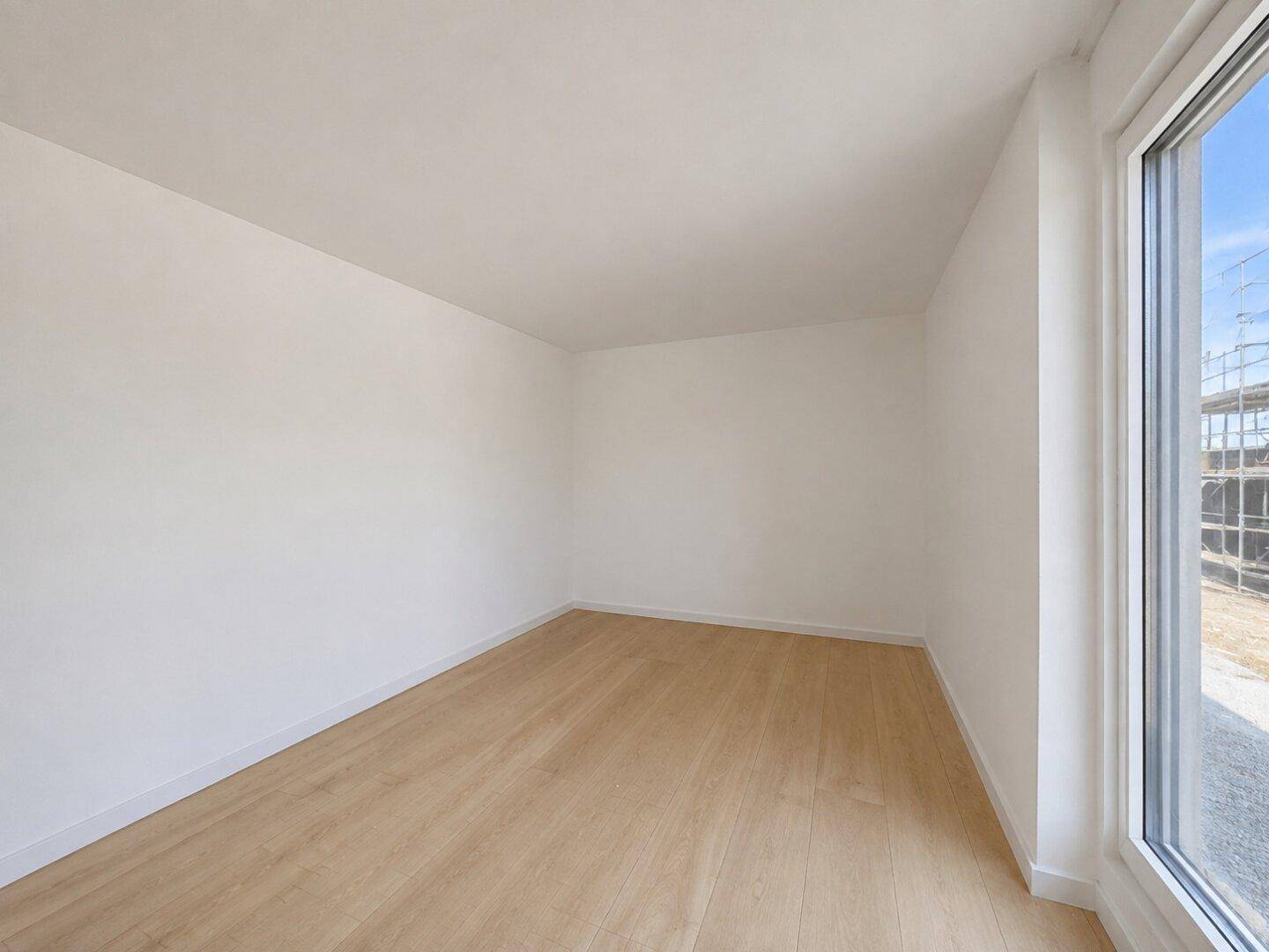 Leeres Schlafzimmer mit hellem Holzboden und großem Fenster, das viel Tageslicht hereinlässt.