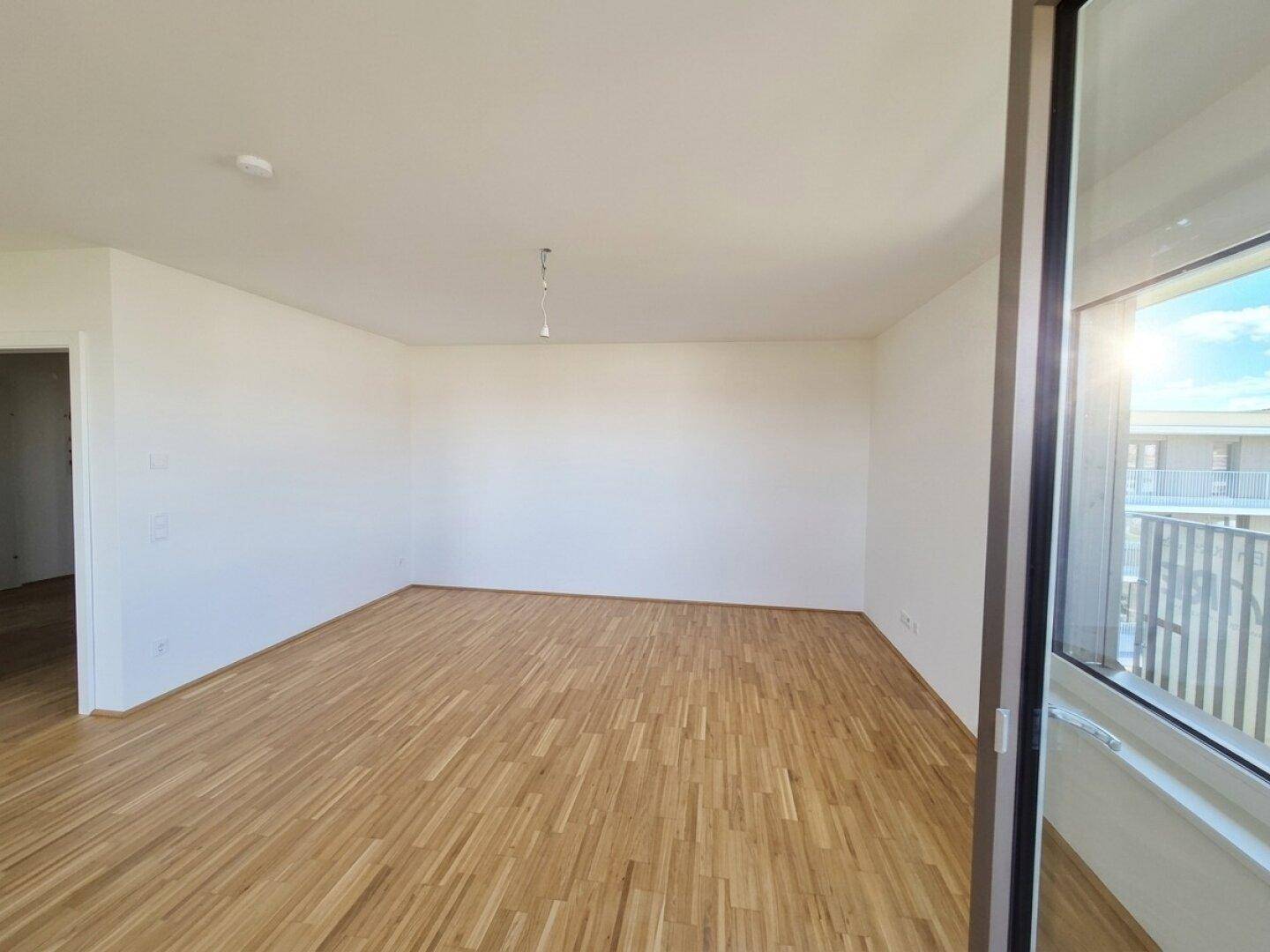 Geräumiges, unmöbliertes Zimmer mit hellem Parkettboden und viel Tageslicht durch die Balkontür.