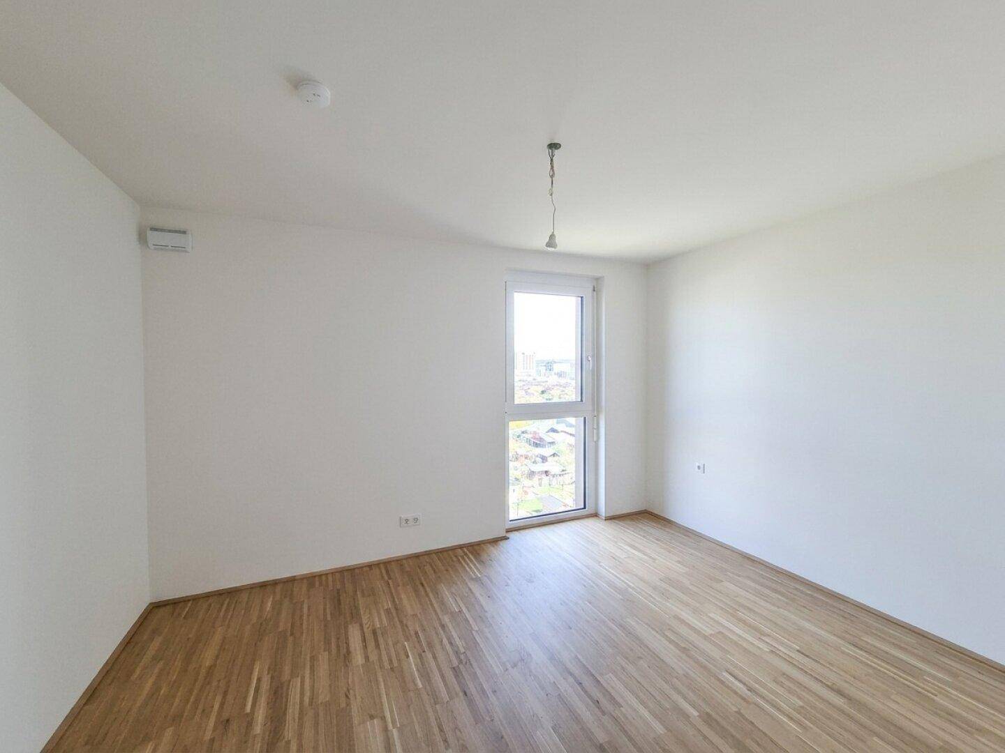 Ruhiges, leeres Schlafzimmer mit Parkettboden und einem Fenster, das den Raum mit Tageslicht füllt.