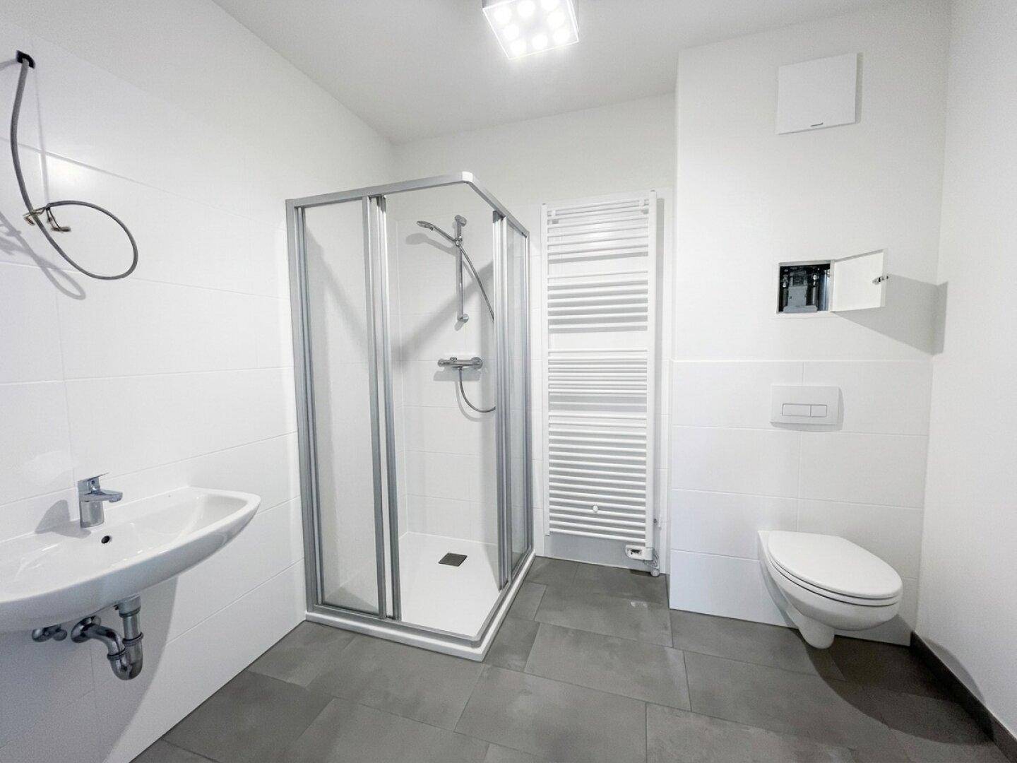 Modernes Badezimmer mit Dusche, Waschbecken, WC und beheiztem Handtuchhalter, in Weiß und Grau gehalten.
