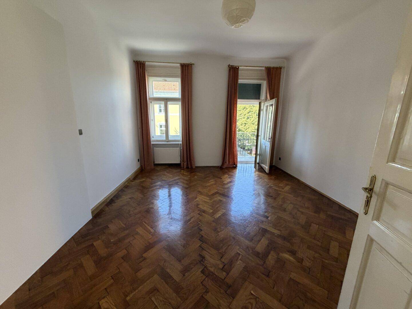 Geräumiges Zimmer mit Fischgrätparkett und Zugang zu einem kleinen Balkon.