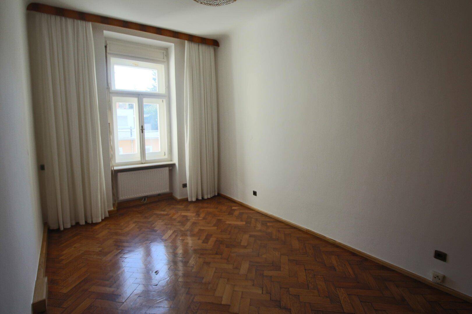 Unmöbliertes Zimmer mit Fischgrätparkett und großem Fenster, ideal als Schlafzimmer.