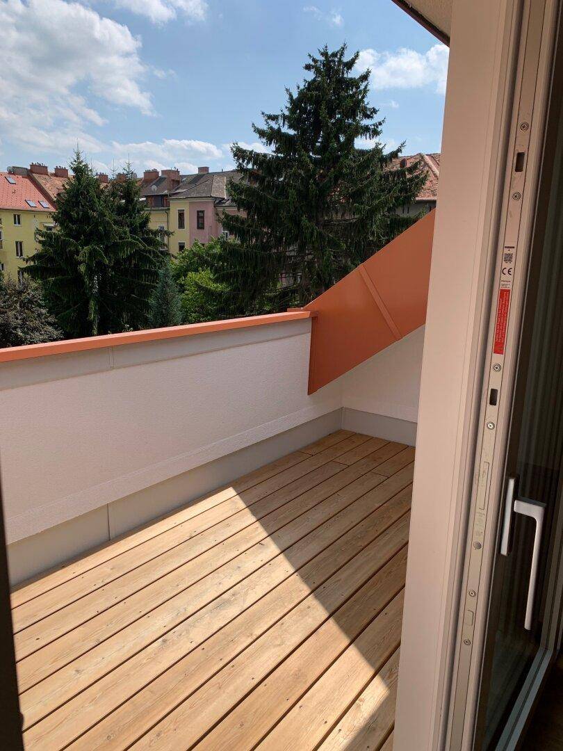 Heller Balkon mit Holzdielen und Blick auf die umliegenden Gebäude und den blauen Himmel.