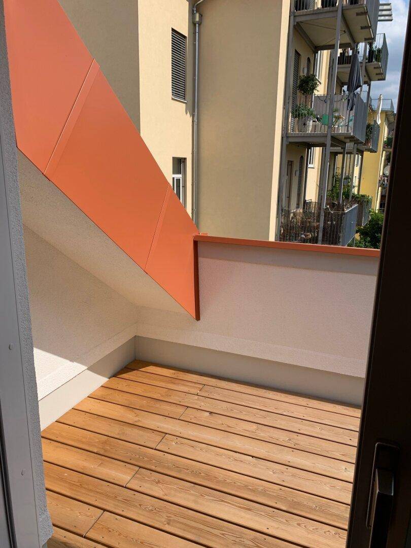 Sonniger Balkon mit Holzboden und Blick auf die benachbarten Gebäude und den klaren Himmel.
