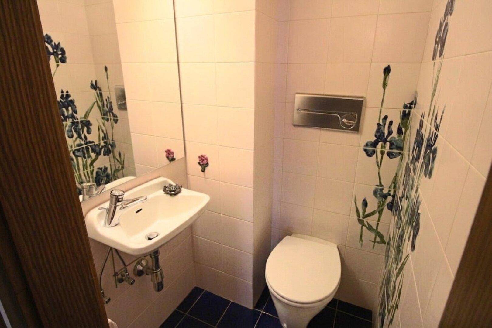 Gäste-WC mit weißem Waschbecken, Toilette und floral gemusterten Fliesen.