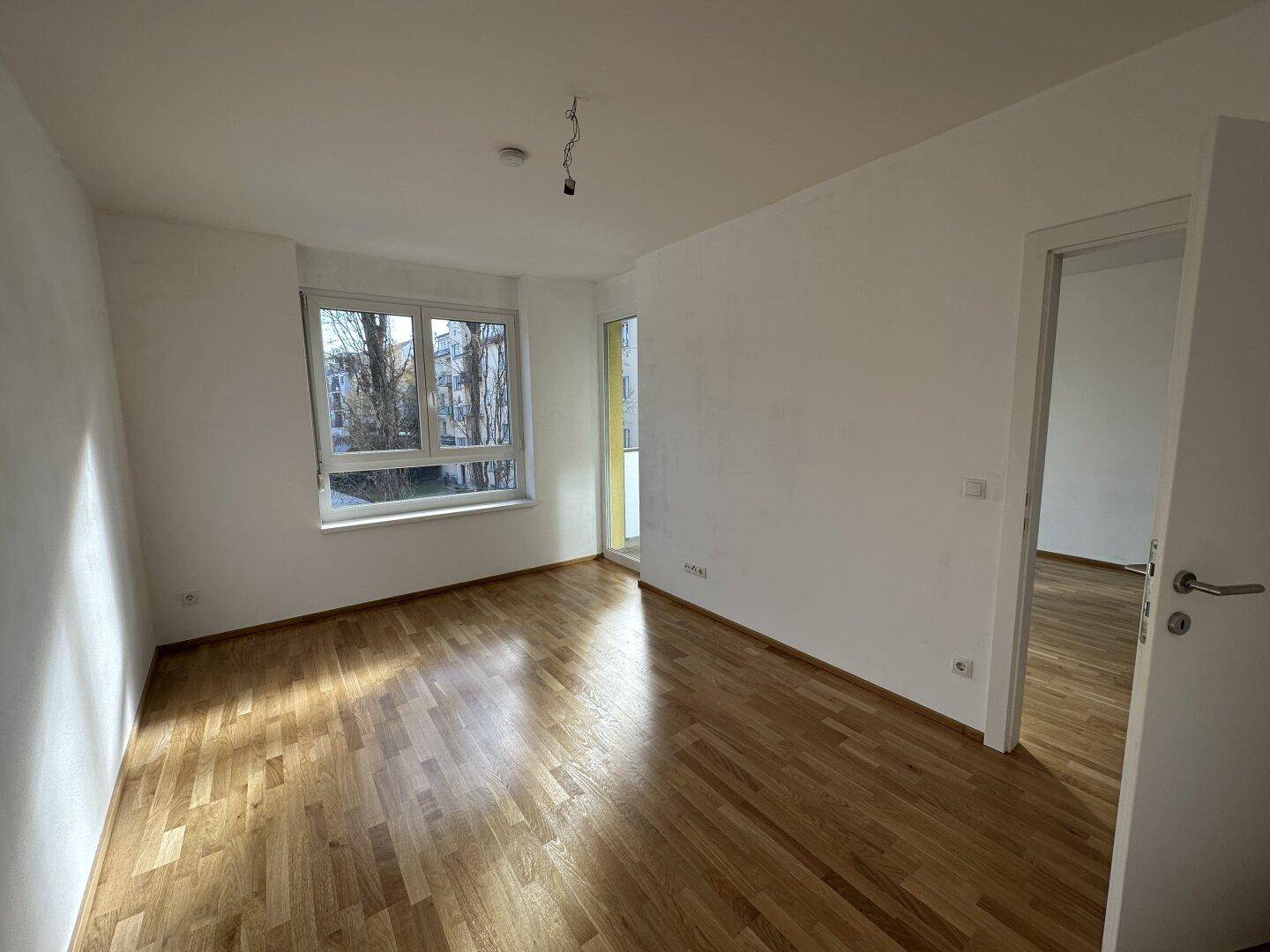 Helles Schlafzimmer mit Parkettboden, weißen Wänden und großem Fenster mit Balkonzugang.