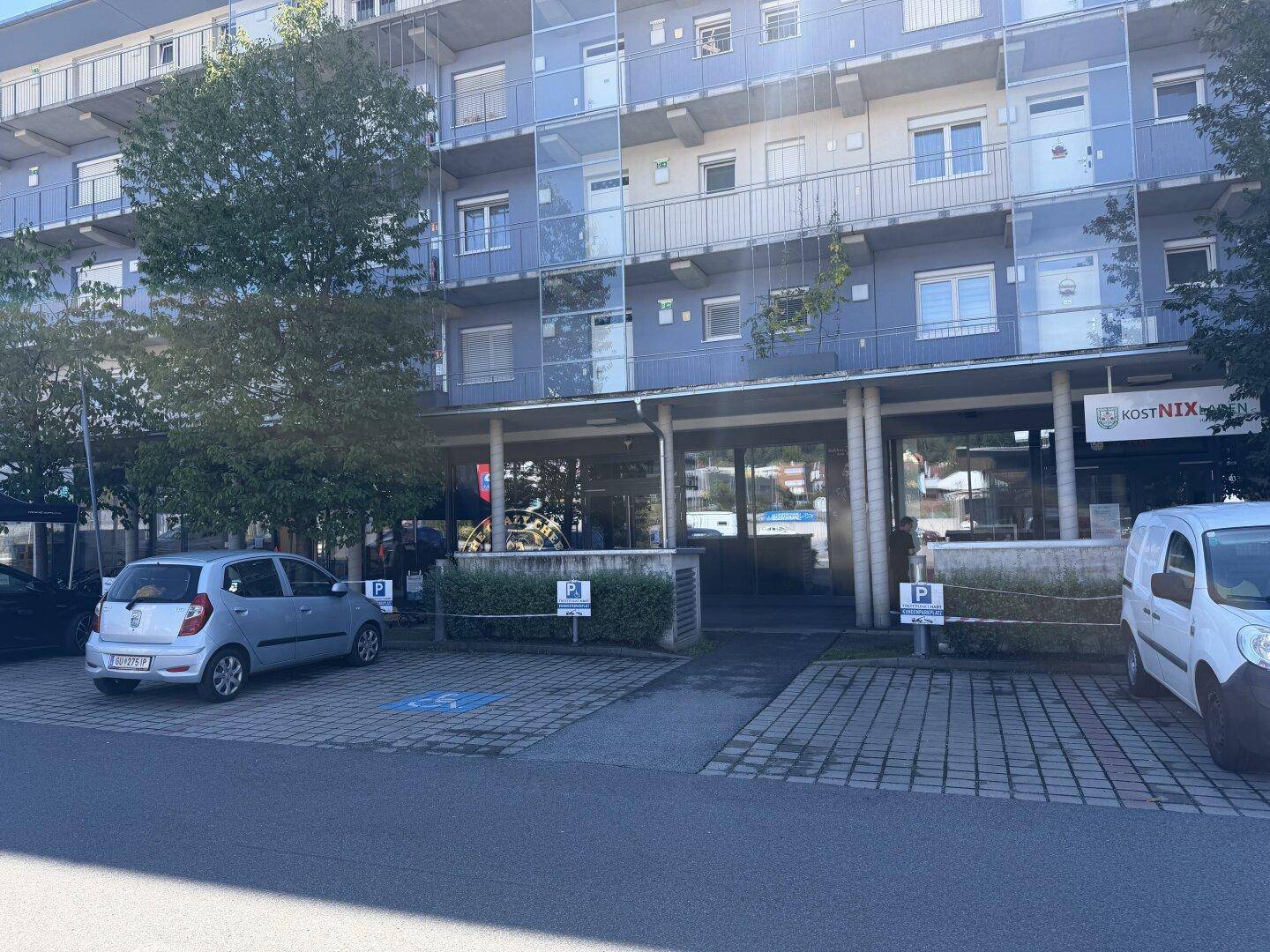 Mehrstöckiges Gebäude mit Parkplätzen davor, umgeben von Bäumen und städtischer Infrastruktur.