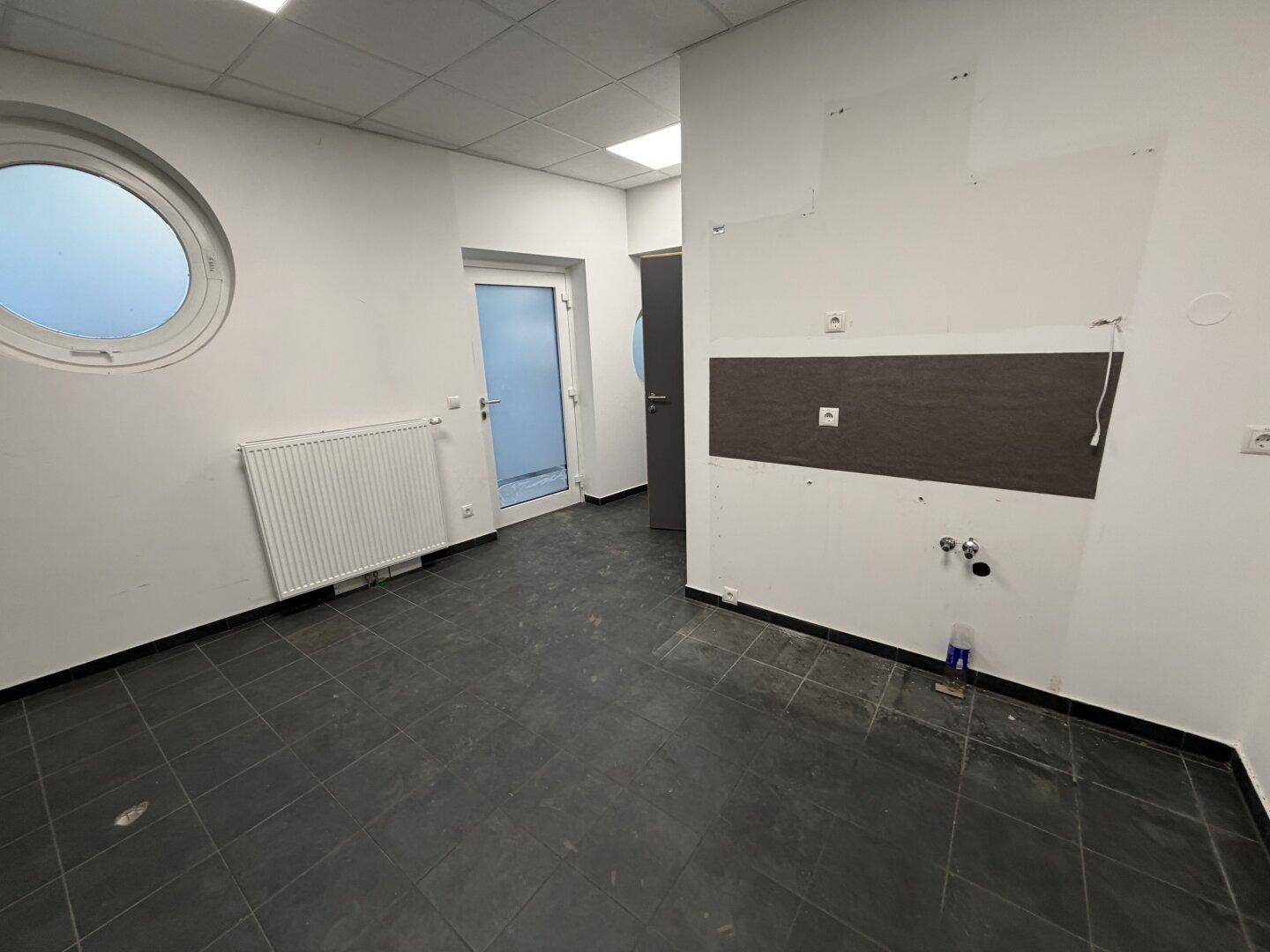 Kleiner Raum mit dunklem Fliesenboden, runden Fenstern und Anschlüssen für eine mögliche Küchenzeile.