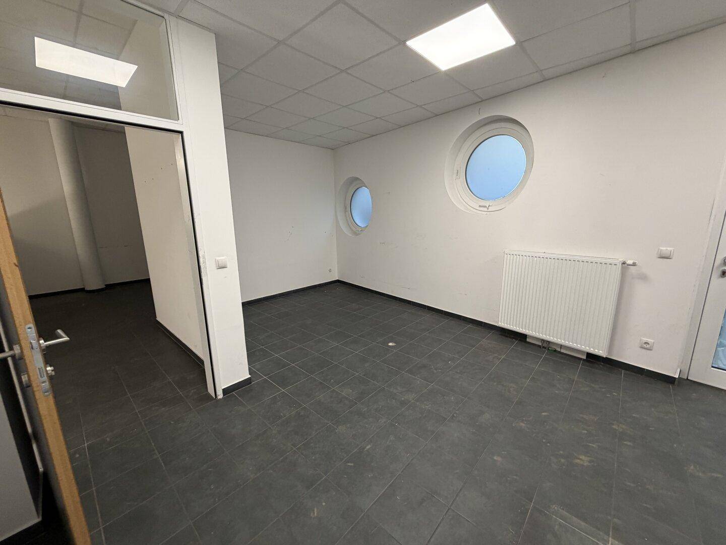 Heller Raum mit dunklem Fliesenboden, weißen Wänden und zwei markanten runden Fenstern.