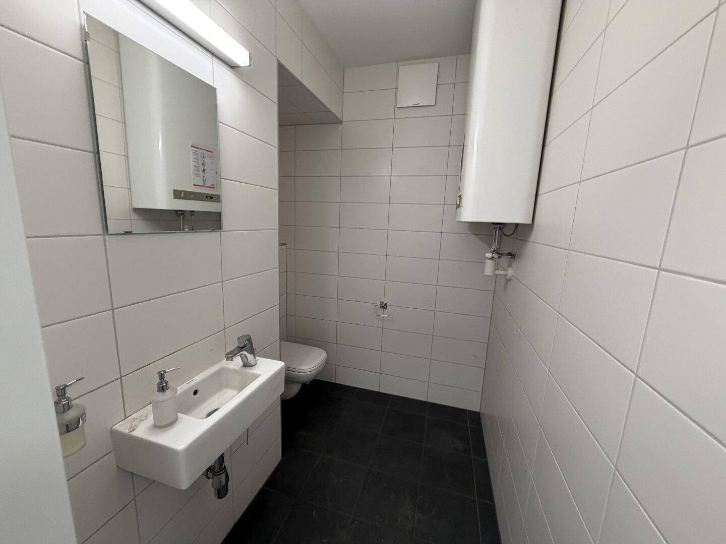 Funktionaler WC-Raum mit weißen Wandfliesen, Waschbecken und Toilette, ausgestattet mit einem Boiler.