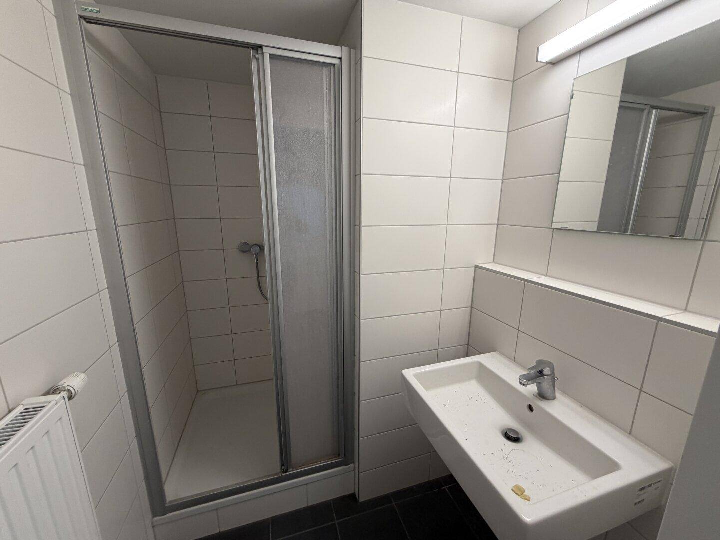 Badezimmer mit weißen Fliesen, Waschbecken und einer Duschkabine, praktisch und hell.