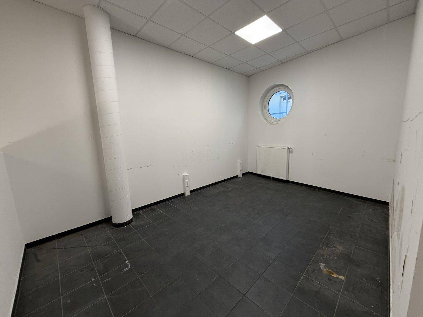 Kleiner, heller Raum mit dunklem Fliesenboden und einem markanten runden Fenster, ideal als Büro.