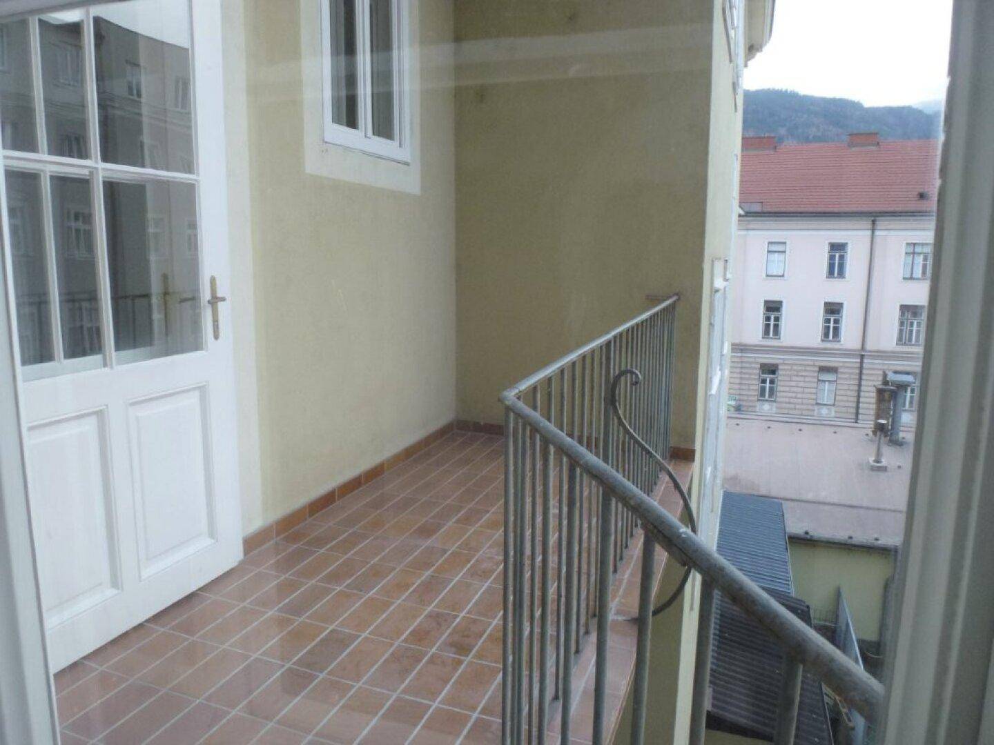 Kleiner Balkon mit Fliesenboden und Metallgeländer, bietet einen Blick auf die umliegenden Gebäude und Berge.