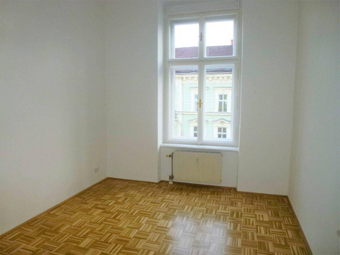 Leeres Zimmer mit hellem Holzparkettboden, weißen Wänden und einem großen Fenster.