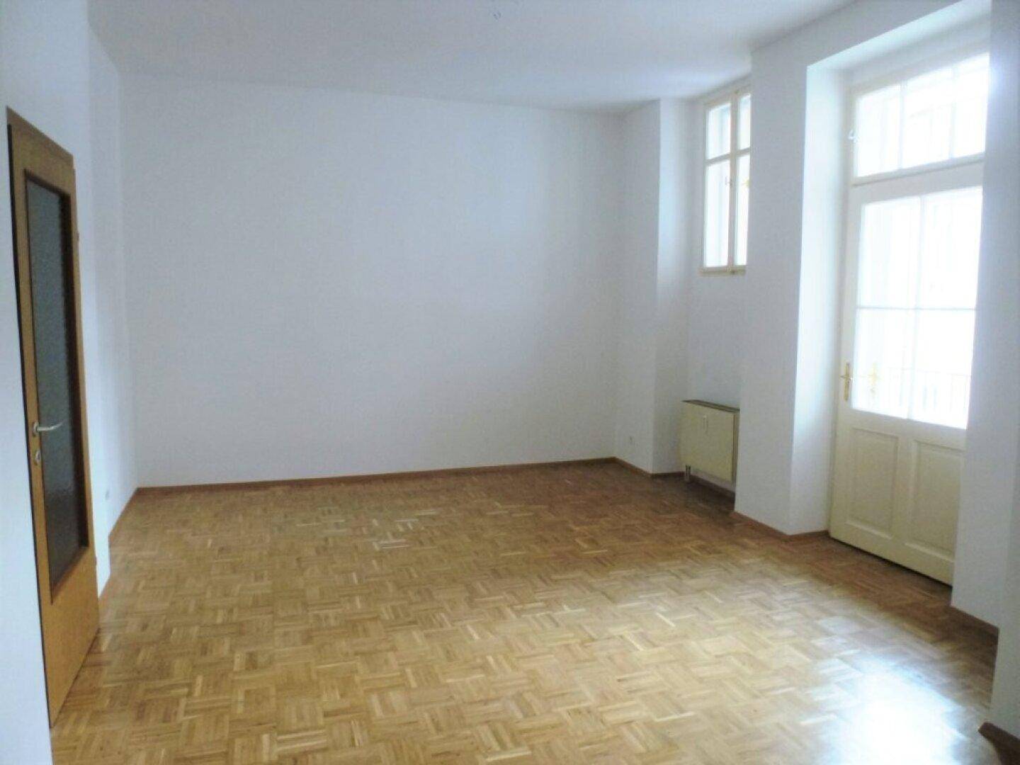 Großes, helles Wohnzimmer mit Holzparkettboden und zwei Fenstern, die viel Tageslicht hereinlassen.