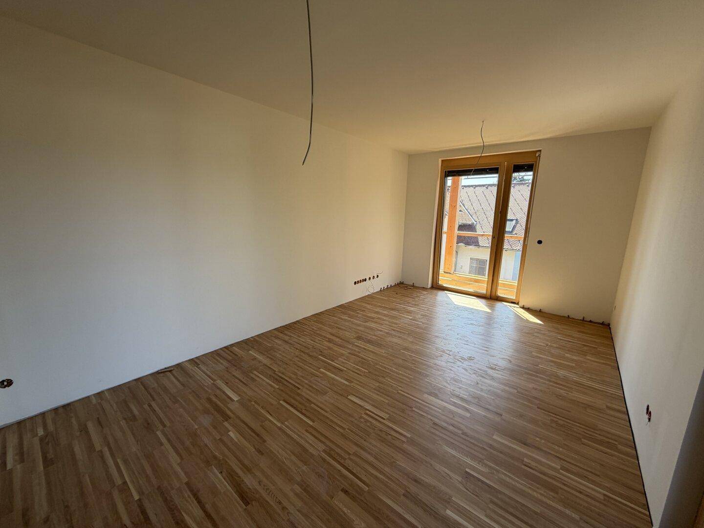 Helles Zimmer mit Parkettboden und Fenster mit Blick auf die umliegenden Dächer und Landschaft.