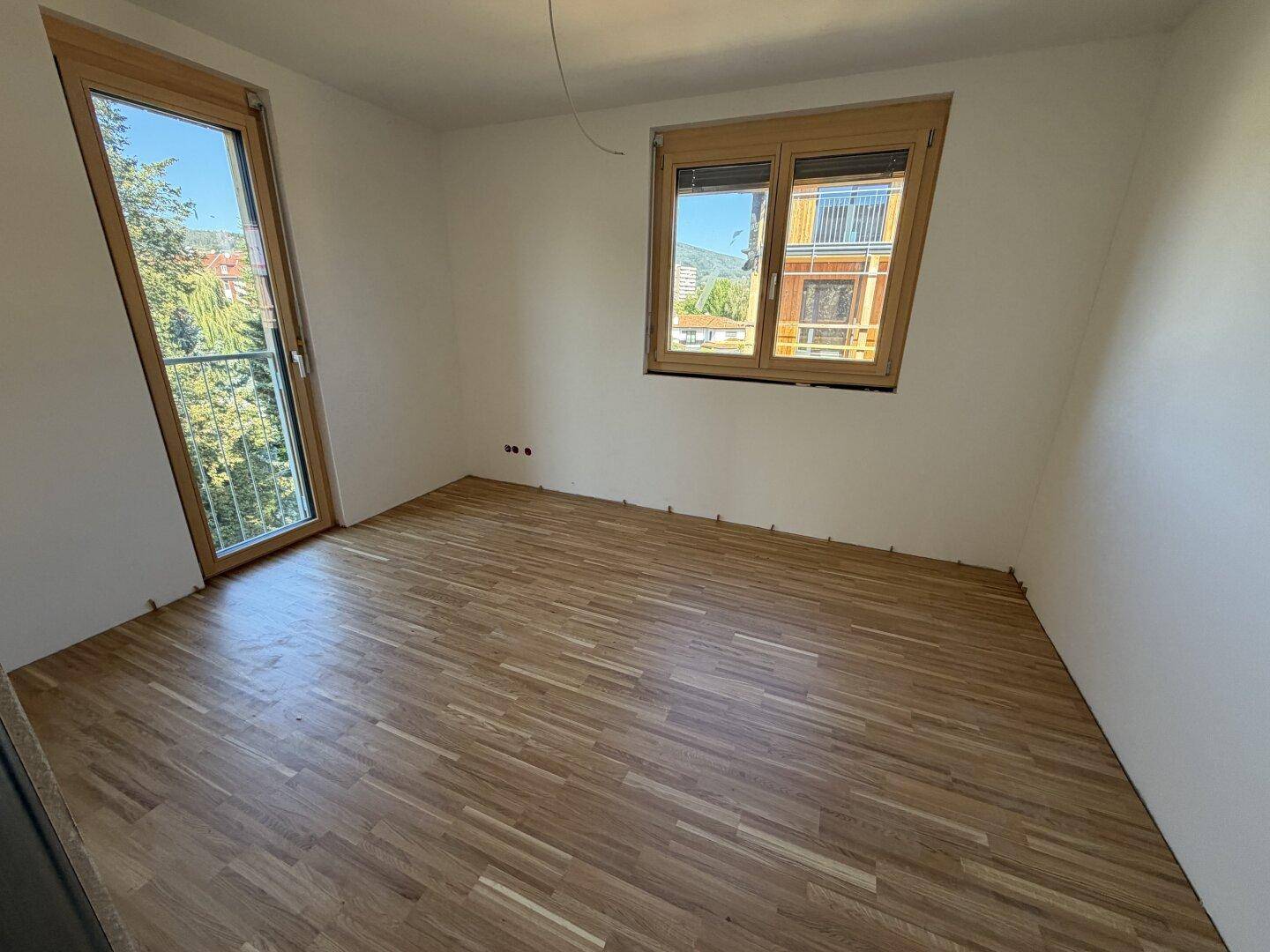 Helles Zimmer mit Parkettboden und zwei Fenstern, eines davon mit französischem Balkon.