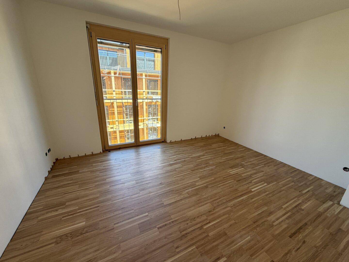 Geräumiges Zimmer mit Parkettboden und großem Fenster mit Blick auf ein Nachbargebäude.