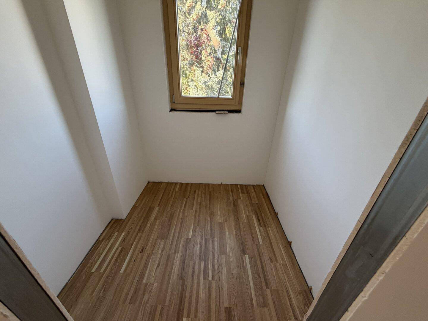 Kleiner Raum mit Parkettboden und Fenster, ideal als Abstellraum oder Ankleidezimmer.