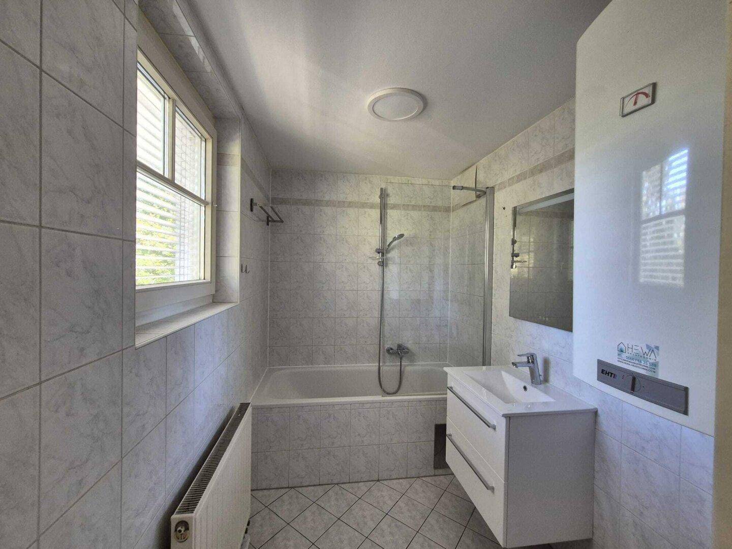 Gefliestes Badezimmer mit Badewanne, Duschfunktion, Einzelwaschbecken und Fenster für Belüftung.