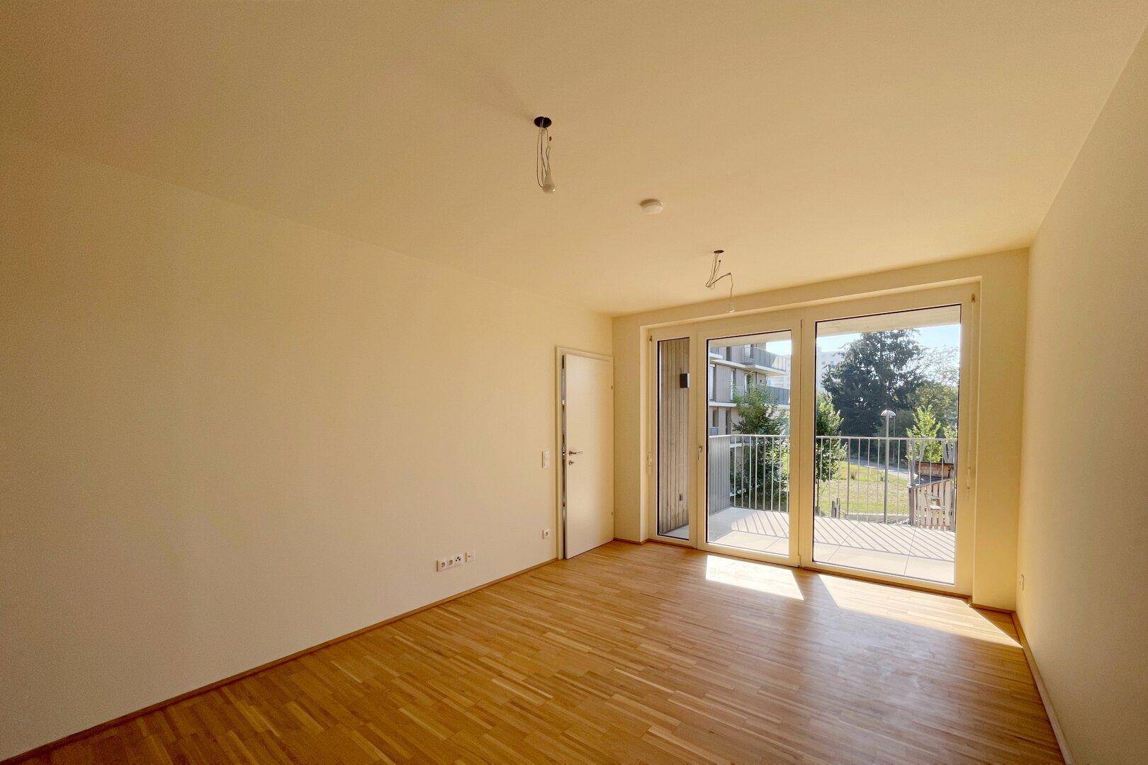 Heller, leerer Raum mit Parkettboden und großen Fenstern, die Zugang zu einem Balkon bieten.