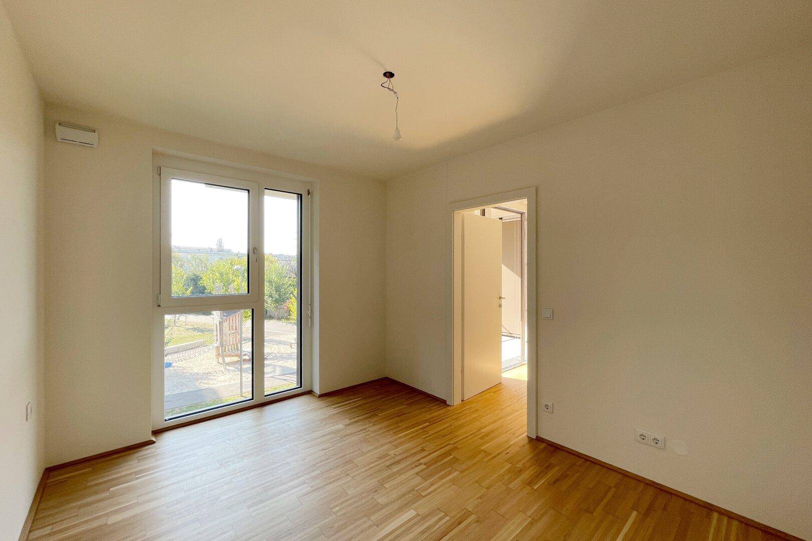 Helles Schlafzimmer mit Parkettboden und großem Fenster, das einen Blick ins Grüne bietet.
