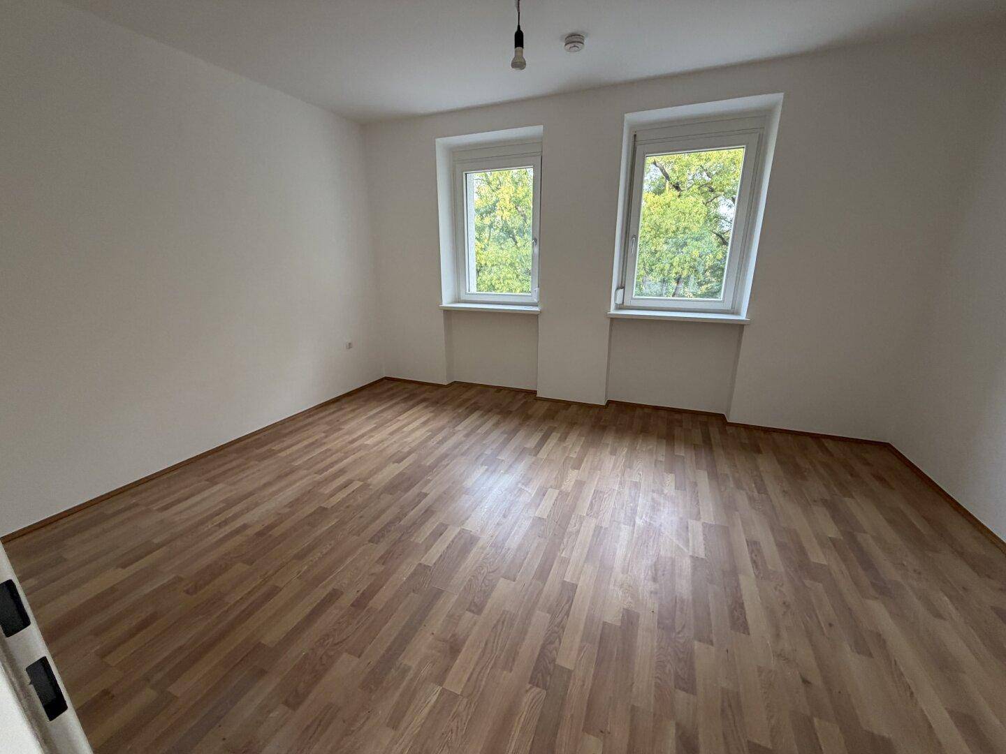 Helles Schlafzimmer mit Laminatboden und zwei Fenstern, die einen Blick ins Grüne bieten.