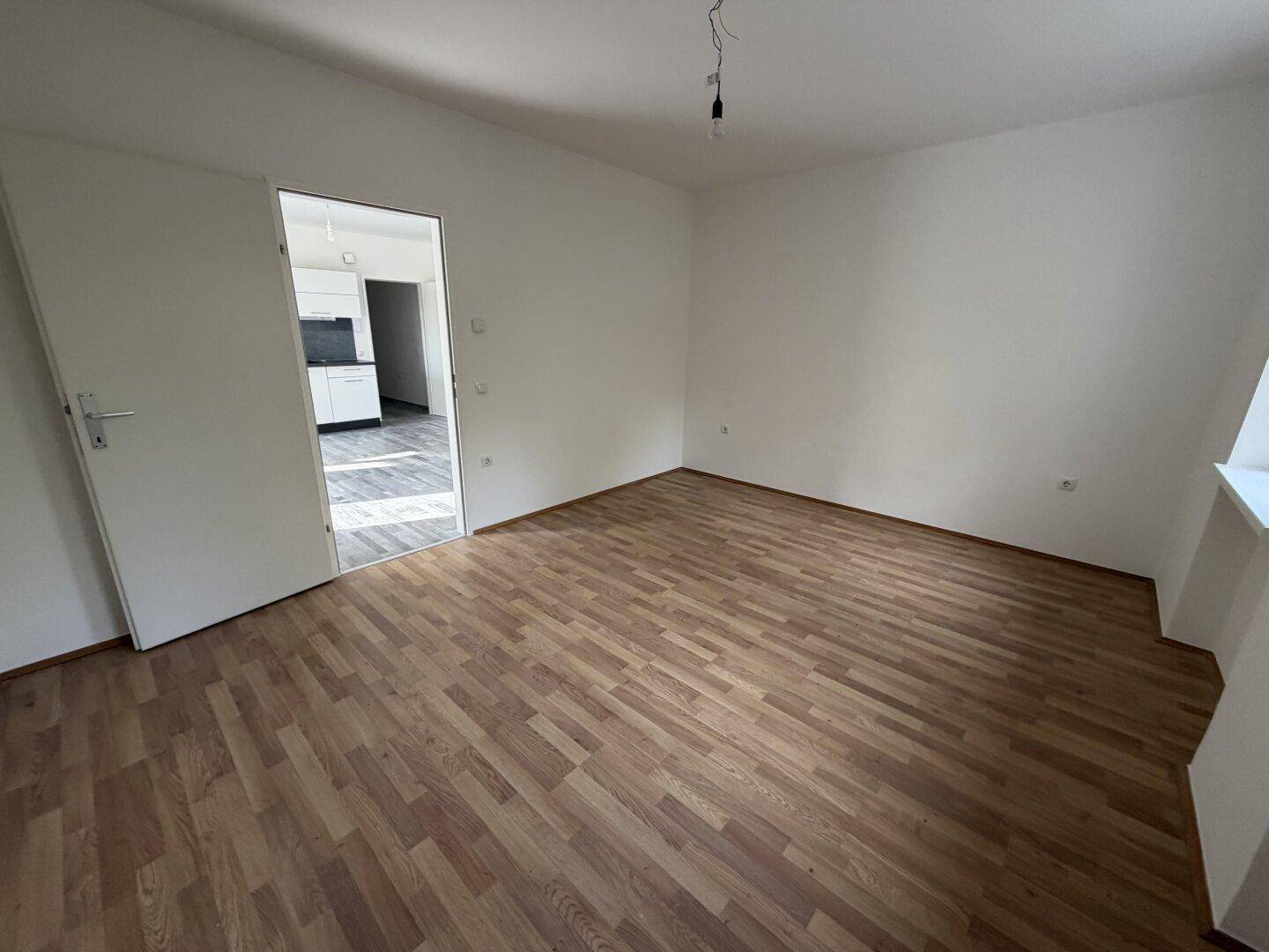Leeres Zimmer mit Laminatboden und offener Tür, die den Blick auf eine moderne Küche freigibt.
