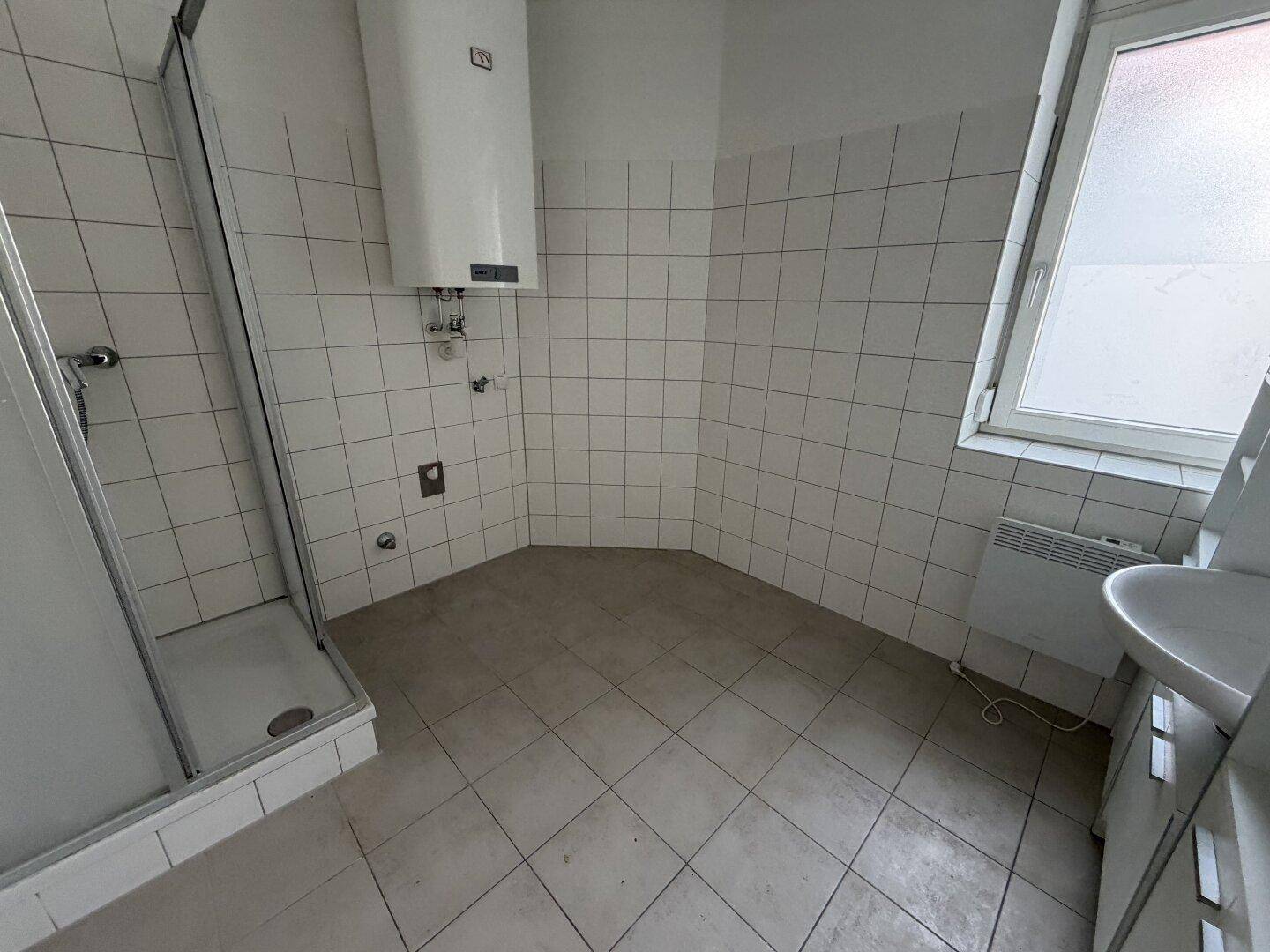 Badezimmer mit weißen Wandfliesen, grauem Fliesenboden, Duschkabine und Fenster.