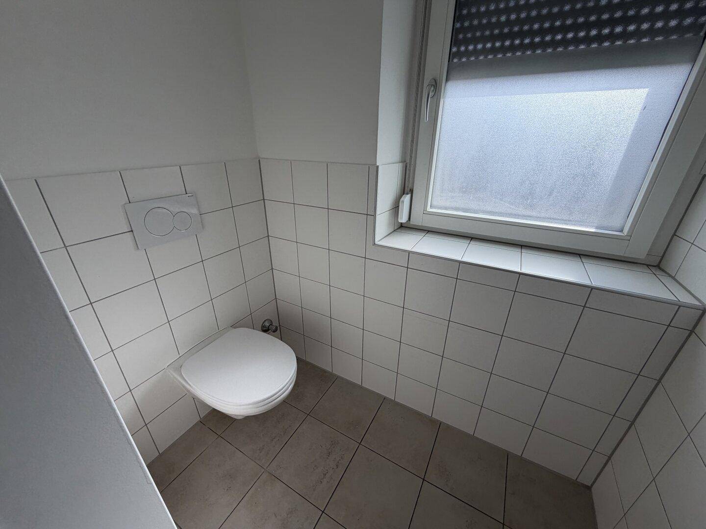 Separates WC mit weißen Fliesen, wandhängendem WC und einem Fenster für Belüftung.