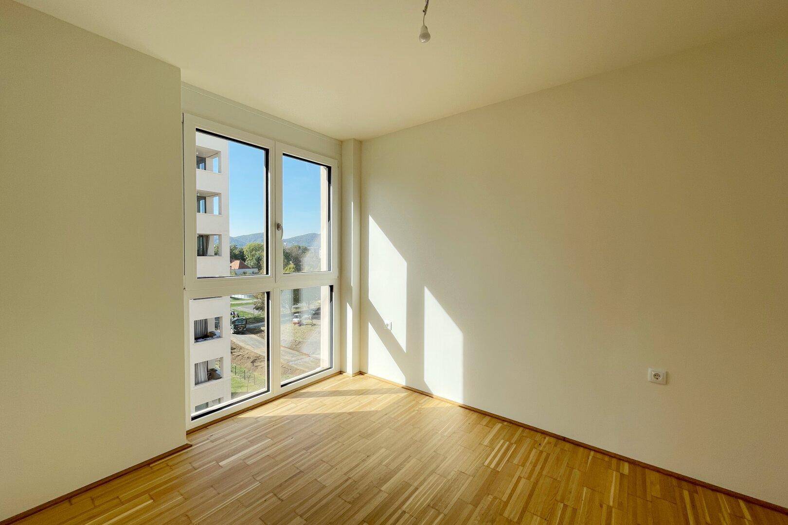 Helles Zimmer mit Parkettboden und großem Fenster mit Blick auf Gebäude und Berge.