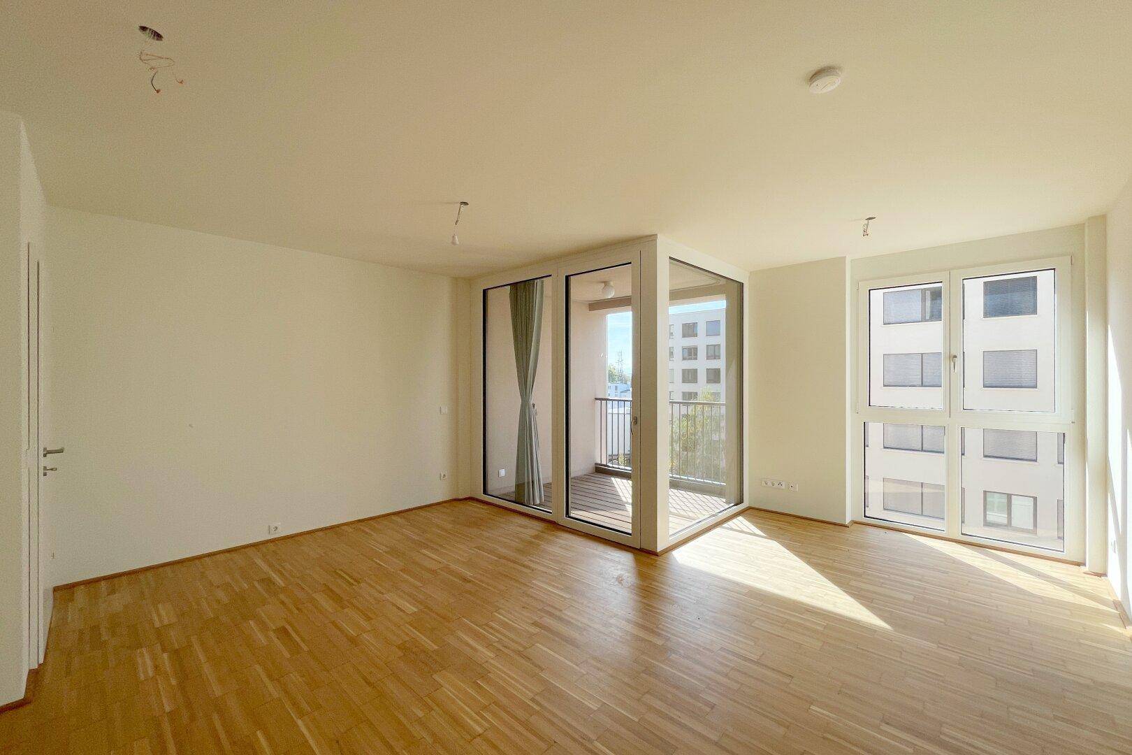 Lichtdurchfluteter Wohnraum mit Parkettboden und Eckfenstern, die Zugang zum Balkon bieten.