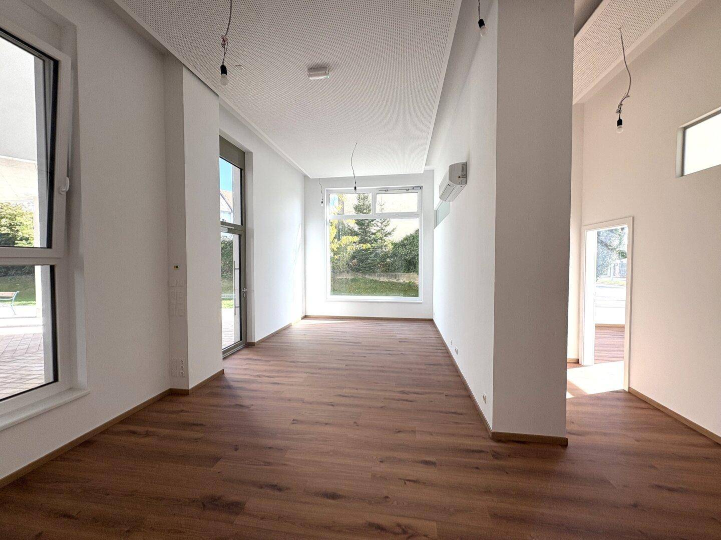 Geräumiger, heller Raum mit Laminatboden, Klimaanlage und großen Fenstern, die viel Tageslicht hereinlassen.