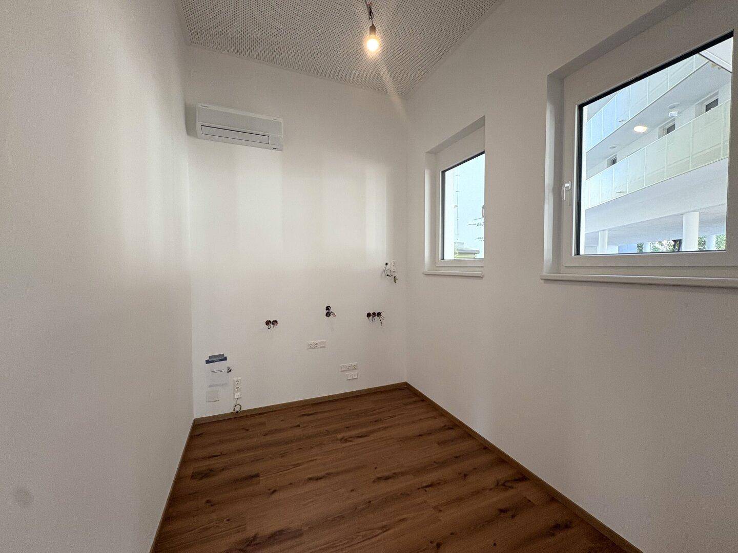 Kleiner Raum mit Laminatboden, Klimaanlage und zwei Fenstern, bereit für die Einrichtung einer Küche oder Nasszelle.
