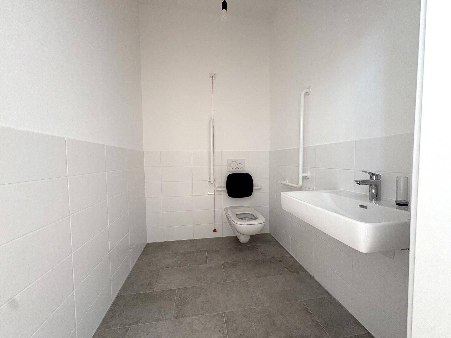 Modernes barrierefreies WC mit weißen Fliesen, grauem Boden, wandhängendem WC und praktischen Haltegriffen.