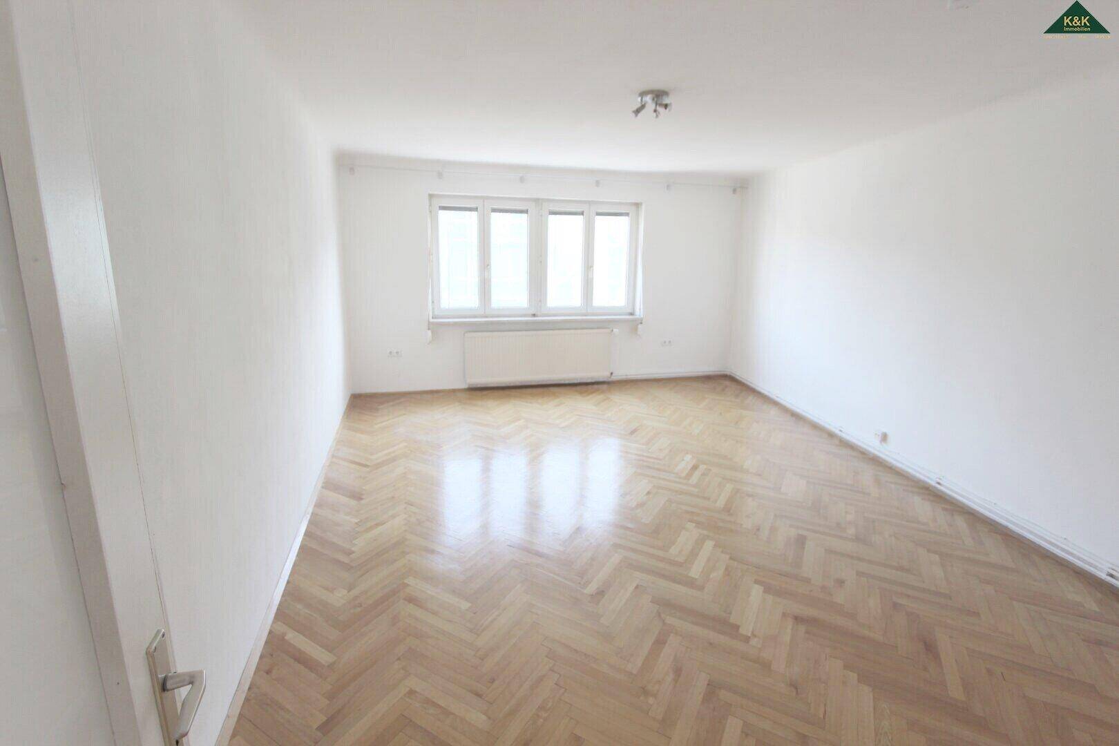Heller, leerer Raum mit weiß gestrichenen Wänden, klassischem Fischgrätparkett und einem großen Fenster.